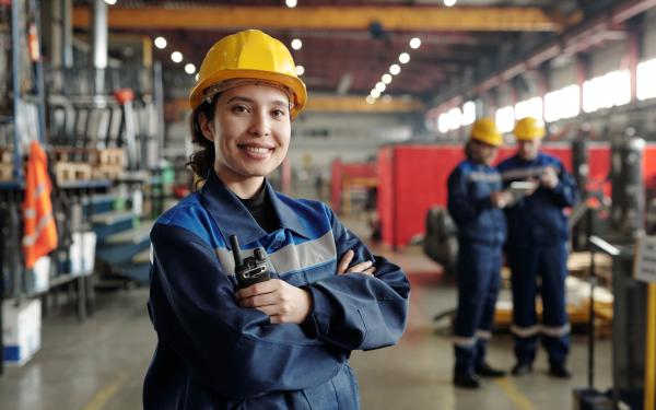 woman skilled migrant standing in a factory with a yellow safety, holding a transceiver and wearing blue jumpsuit